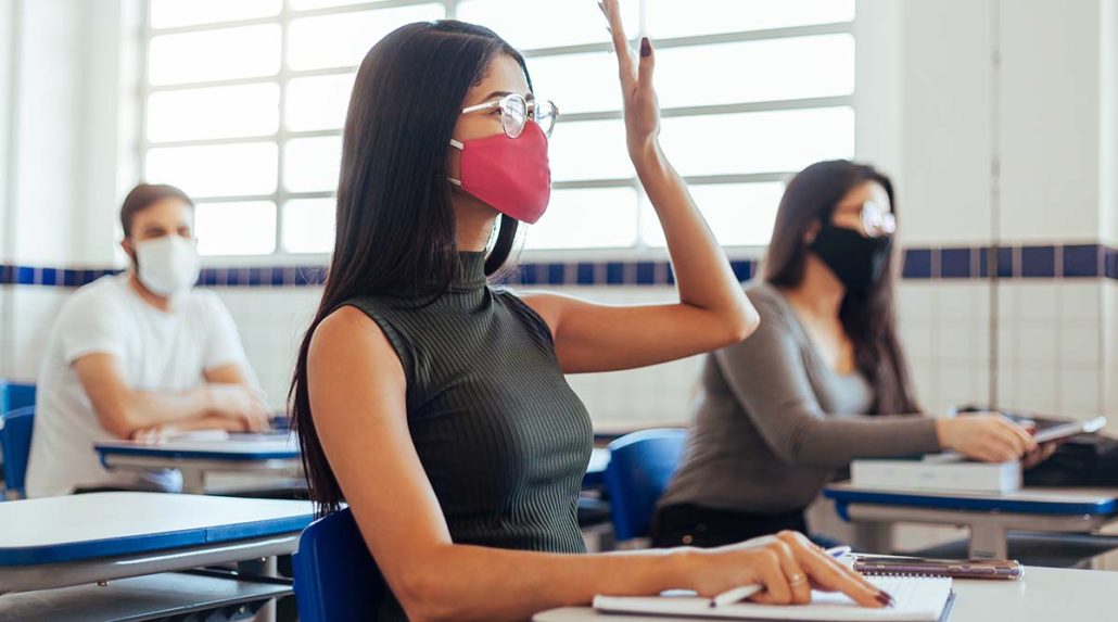 A female student raising her hand with a mask on in a classroom, participating in hybrid learning.
